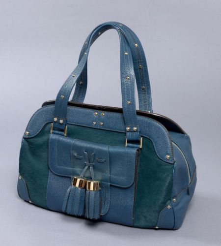 LUELLA, grand sac porté main en cuir grainé bleu canard et poulain à la couleur. Fermeture zippée. Poche à rabat sur le devant ornée de deux gros pompons, une poche zippée au dos. Garniture cloutée en métal doré. Intérieur en daim beige, une grande poche 